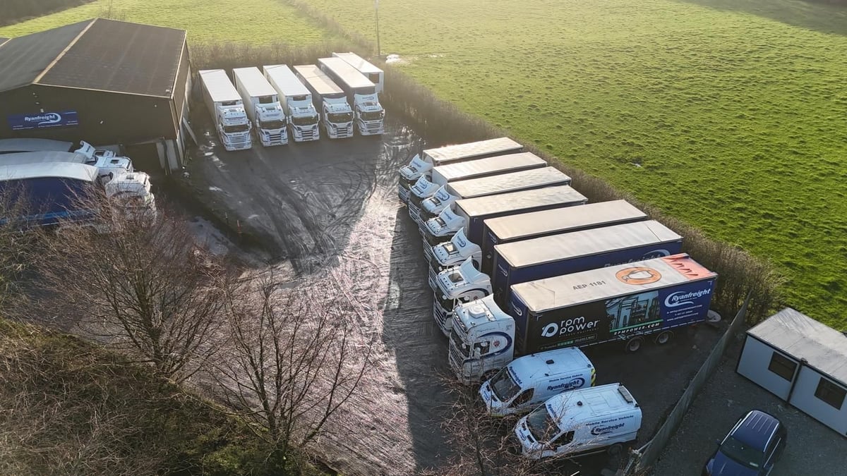 Aerial view of commercial trucks and trailers parked in a row on a gravel area next to a green field