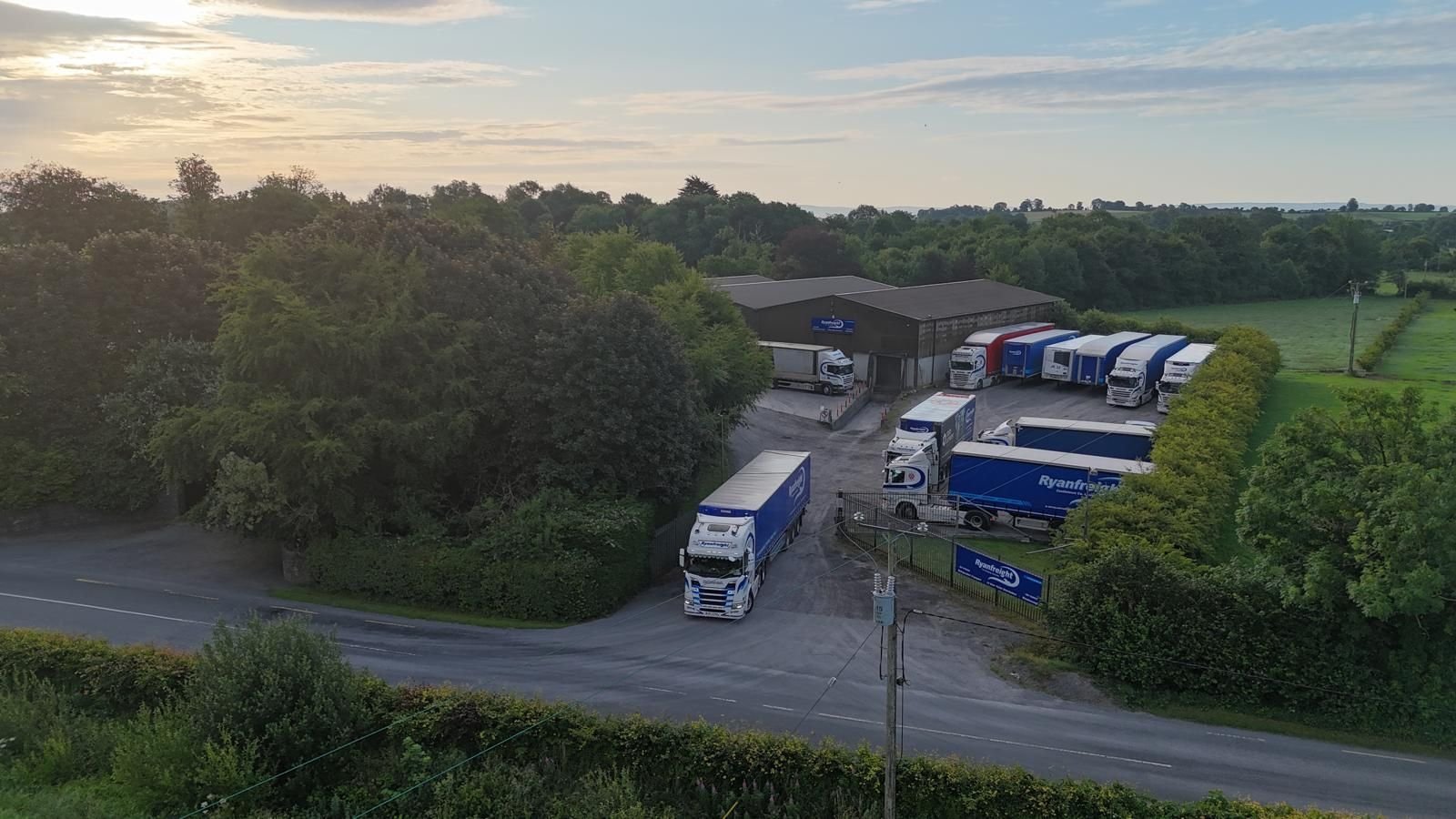 Aerial view of an industrial facility with trucks parked outside, surrounded by green trees and countryside landscape at dusk